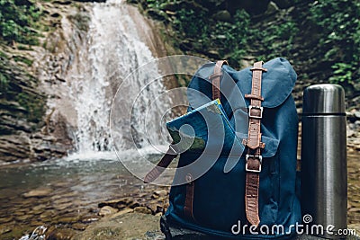 Hipster Blue Backpack, Map And Thermos Closeup. View From Front Tourist Traveler Bag On Waterfall Background. Adventure Hiking Con Stock Photo