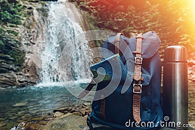 Hipster Blue Backpack, Map And Thermos Closeup. View From Front Tourist Traveler Bag On Waterfall Background. Adventure Hiking Con Stock Photo