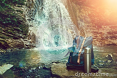 Hipster Blue Backpack, Map And Thermos. View From Front Tourist Traveler Bag On Waterfall Background. Wanderlust Adventure Hiking Stock Photo