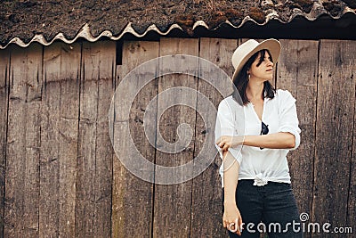 Hipster girl in white casual outfit posing on background of old wooden cabin in mountains. Stylish woman in hat standing near old Stock Photo