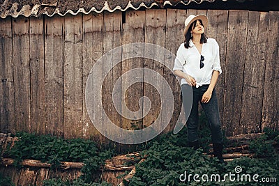Hipster girl in white casual outfit posing on background of old wooden cabin in mountains. Stylish woman in hat standing near old Stock Photo