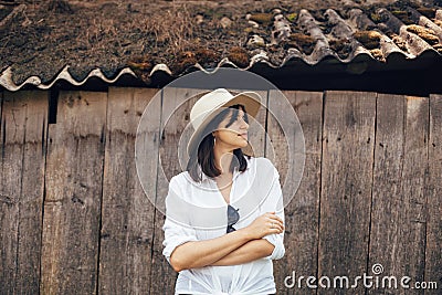 Hipster girl in white casual outfit posing on background of old wooden cabin in mountains. Stylish woman in hat standing near old Stock Photo