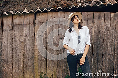 Hipster girl in white casual outfit posing on background of old wooden cabin in mountains. Stylish woman in hat standing near old Stock Photo