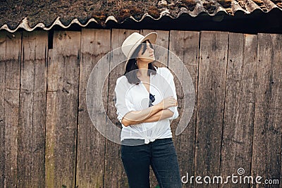 Hipster girl in white casual outfit posing on background of old wooden cabin in mountains. Stylish woman in hat standing near old Stock Photo