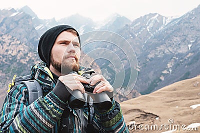 A hipster man with a beard in a hat, a jacket, and a backpack in the mountains holds binoculars, adventure, tourism Stock Photo