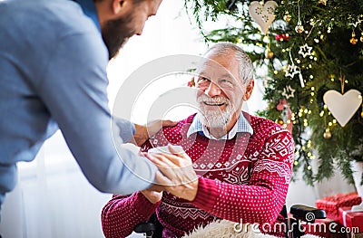 A hipster man giving drink to his senior father in wheelchair at Christmas time. Stock Photo