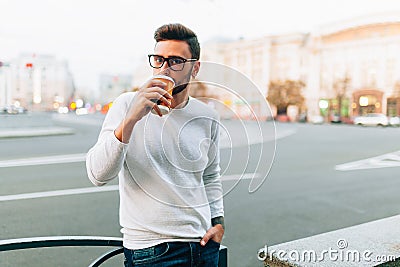 Hipster man standing with takeaway coffee, smiling plesantly, walking on th city street. Happy carefree handsome guy in eyewear Stock Photo