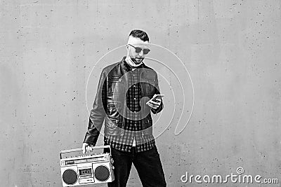 Hipster man using smartphone while listening music with a vintage boombox player against a wall background - Man wearing open face Stock Photo