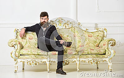 Hipster on strict face sits alone. Rich and lonely concept. Owner of luxury apartment sits on sofa, white wall Stock Photo