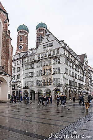 Feb 2, 2020 - Munich, Germany: HIRMER House on street of Munchen with domed towers of Gotchic church Frauenkirche Editorial Stock Photo