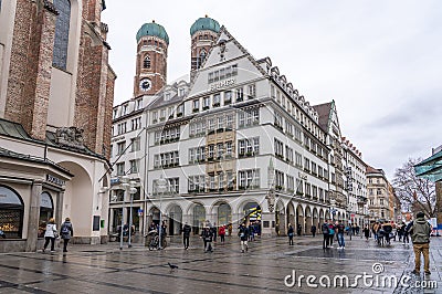 Feb 2, 2020 - Munich, Germany: HIRMER House on street of Munchen with domed towers of Gotchic church Frauenkirche Editorial Stock Photo