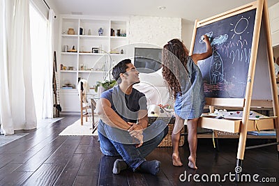 Hispanic dad sitting on the floor in sitting room watching his young daughter drawing on blackboard Stock Photo