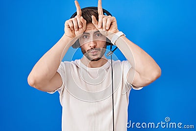 Hispanic man with beard listening to music wearing headphones doing funny gesture with finger over head as bull horns Stock Photo