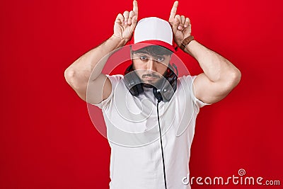 Hispanic man with beard wearing gamer hat and headphones doing funny gesture with finger over head as bull horns Stock Photo