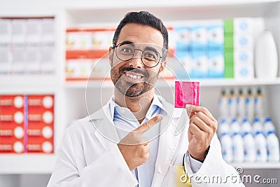 Hispanic man with beard working at pharmacy drugstore holding condom smiling happy pointing with hand and finger Stock Photo