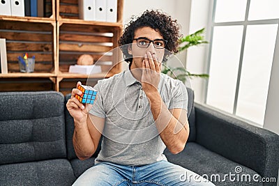 Hispanic man with curly hair playing colorful puzzle cube intelligence game covering mouth with hand, shocked and afraid for Editorial Stock Photo