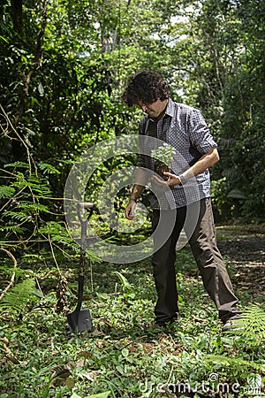 Hispanic man planting a small plant in a green field surrounded by trees during a sunny day Stock Photo