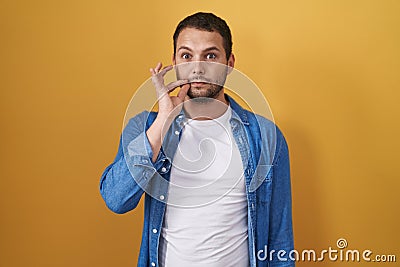 Hispanic man standing over yellow background mouth and lips shut as zip with fingers Stock Photo