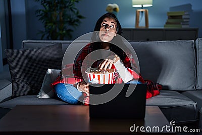 Hispanic woman eating popcorn watching a movie on the sofa making fish face with lips, crazy and comical gesture Stock Photo