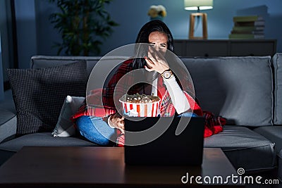 Hispanic woman eating popcorn watching a movie on the sofa smelling something stinky and disgusting, intolerable smell, holding Stock Photo