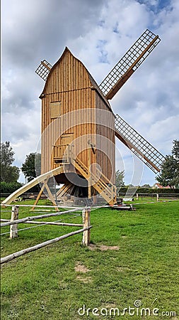 Historic windmill made of light brown wood . Wooden building with barrier band , green grass and very white cloudy sky . Ancient Stock Photo