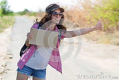 Hitchhiking woman travel. Beautiful young female hitchhiker in a cap and sunglasses smiling while standing by the road during Stock Photo