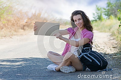 Hitchhiking woman travel. Happy young woman is sitting on the road with a tourist backpack and holding a cardboard in Stock Photo