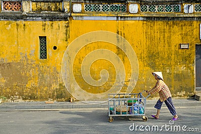 Hoi An / Vietnam, 12/11/2017: Local Vietnamese woman with rice hat pushing a carriage in front of traditional houses with Editorial Stock Photo