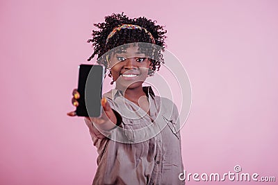 Holding black phone in hand. Attractive afro american woman in casual clothes at pink background in the studio Stock Photo