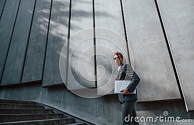 Holds laptop in hands. Young businessman in grey formal wear is outdoors in the city Stock Photo