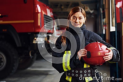 Holds red hat in hands. Female firefighter in protective uniform standing near truck Stock Photo