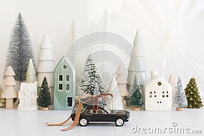 Holidays are coming! Stylish little car carrying christmas tree on background of Christmas miniature snowy village. Merry Stock Photo
