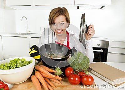 Home cook woman in red apron at domestic kitchen holding cooking pot with hot soup smelling vegetable stew Stock Photo