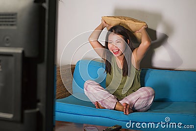 Home lifestyle portrait of young pretty and happy Asian Chinese woman laughing watching korean comedy TV show sitting at on living Stock Photo