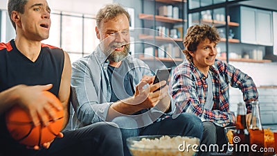 At Home Three Fans Sitting on Couch Watch Basketball Game on TV, Use Smartphone App for Checking Stock Photo