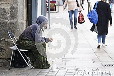 Homeless beggar sat on busy street wearing a hoodie with cup for change in the UK with shoppers in the background Editorial Stock Photo