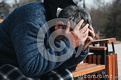 homeless elderly old Caucasian man with depression holds head in hands while sitting on park bench Stock Photo
