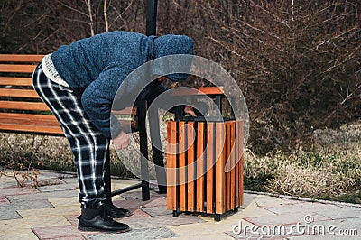 homeless elderly old Caucasian man rummages for food and garbage in trash can in a park in autumn Stock Photo