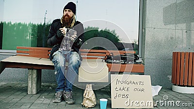 Homeless and jobless european man with cardboard sign eat sandwich on bench at city street because of immigrants crisis Stock Photo