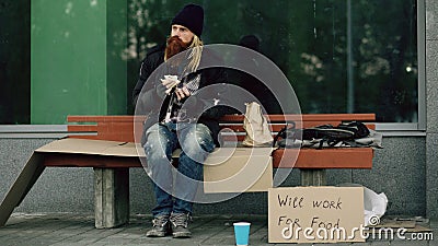 Homeless and jobless european man with cardboard sign eat sandwich on bench at city street because of immigrants crisis Stock Photo