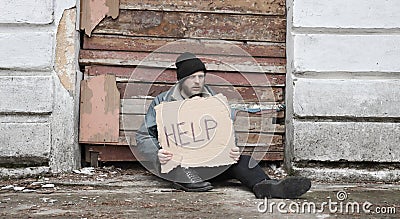 Homeless man sits and holds piece of cardboard with inscription Help. Refugee looking for job. Male tramp in dirty clothes, hat Stock Photo
