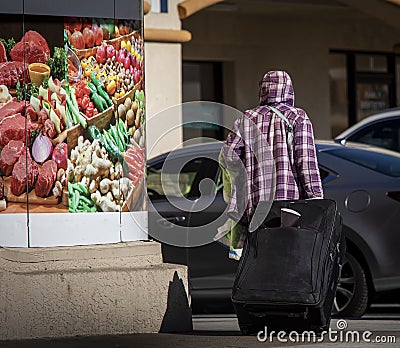 Homeless person pulling suitcase passing a market sign with a photo of various Stock Photo