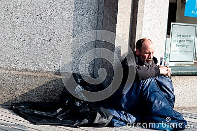 A homeless person siting down holds a cup begging for money while covered in a sleeping bag Editorial Stock Photo