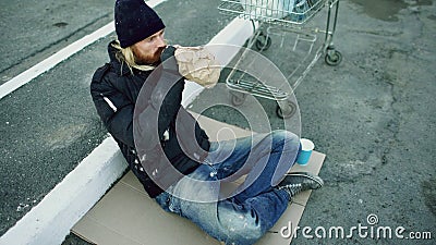 Homeless young man in dirty clothes drink alcohol sitting near shopping cart on the street at cold winter day Stock Photo