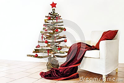 Homemade alternative Christmas tree of rustic raw wood branches decorated with fairy lights and red balls next to an armchair with Stock Photo