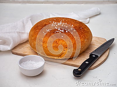 Homemade bread on a wooden unpainted board with coarse salt in a white salt shaker and a kitchen knife, next to it is a white Stock Photo
