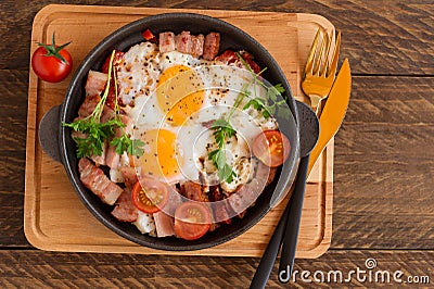 Homemade fried eggs with bacon and tomatoes in a frying pan on wooden background. Classic breakfast. Top view Stock Photo