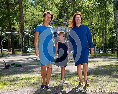 Homosexual lesbian couple hold daughter`s hands. A girl walks with two mothers in the park. Two married women and a Stock Photo