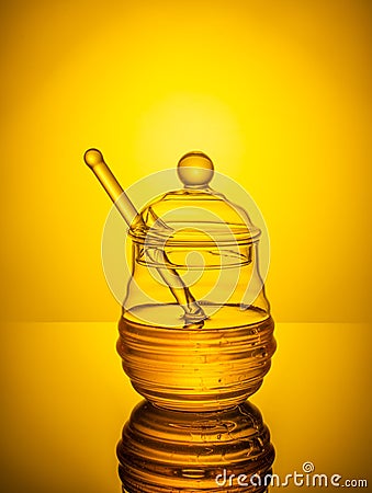 Honey in a glass jar with a stick on a yellow background. A jar of honey is reflected off the surface of the table. Stock Photo