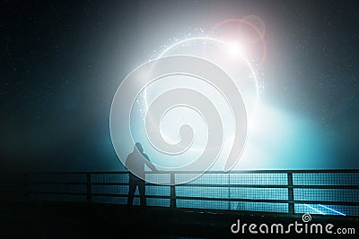 A hooded figure, standing with back to camera on a bridge, looking at a glowing, mystic, science fiction portal. On a foggy night Stock Photo